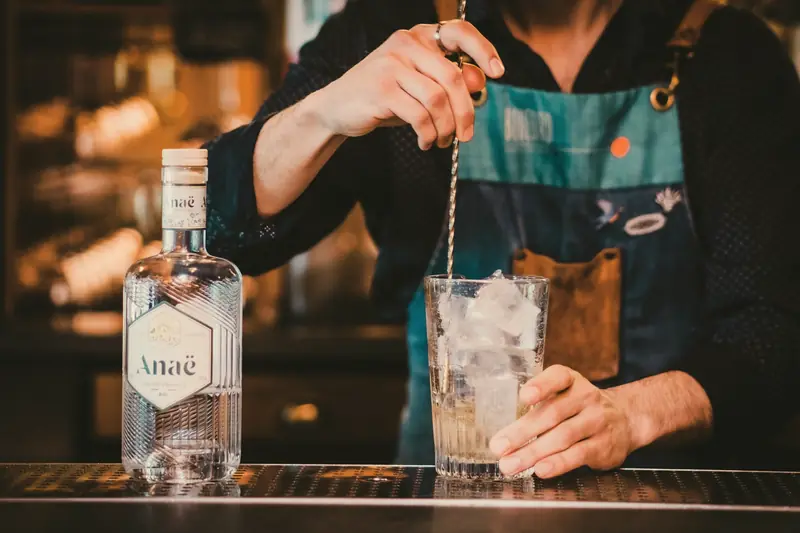 Bartender de avental mexendo um drink com colher bailarina ao lado de uma garrafa de gin